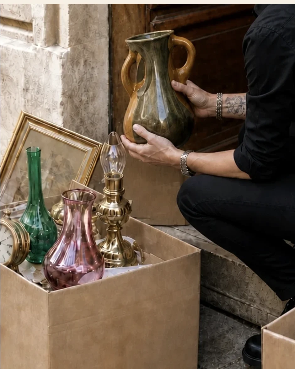 Stéfani soulève un pichet ancien au-dessus de cartons remplis de vases et de lampes, prêts pour le dépôt-vente